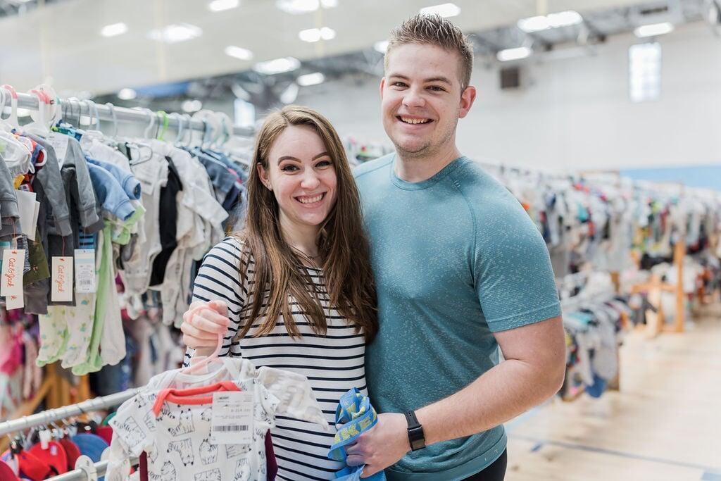 An expecting mom holding some onesies with her husband at a JBF sale.
